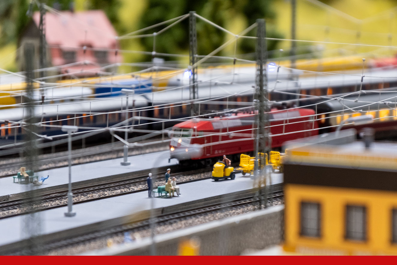 Modellbahnszene mit rotem Triebzug auf Gleisen, Oberleitungen und Figuren auf dem Bahnsteig
