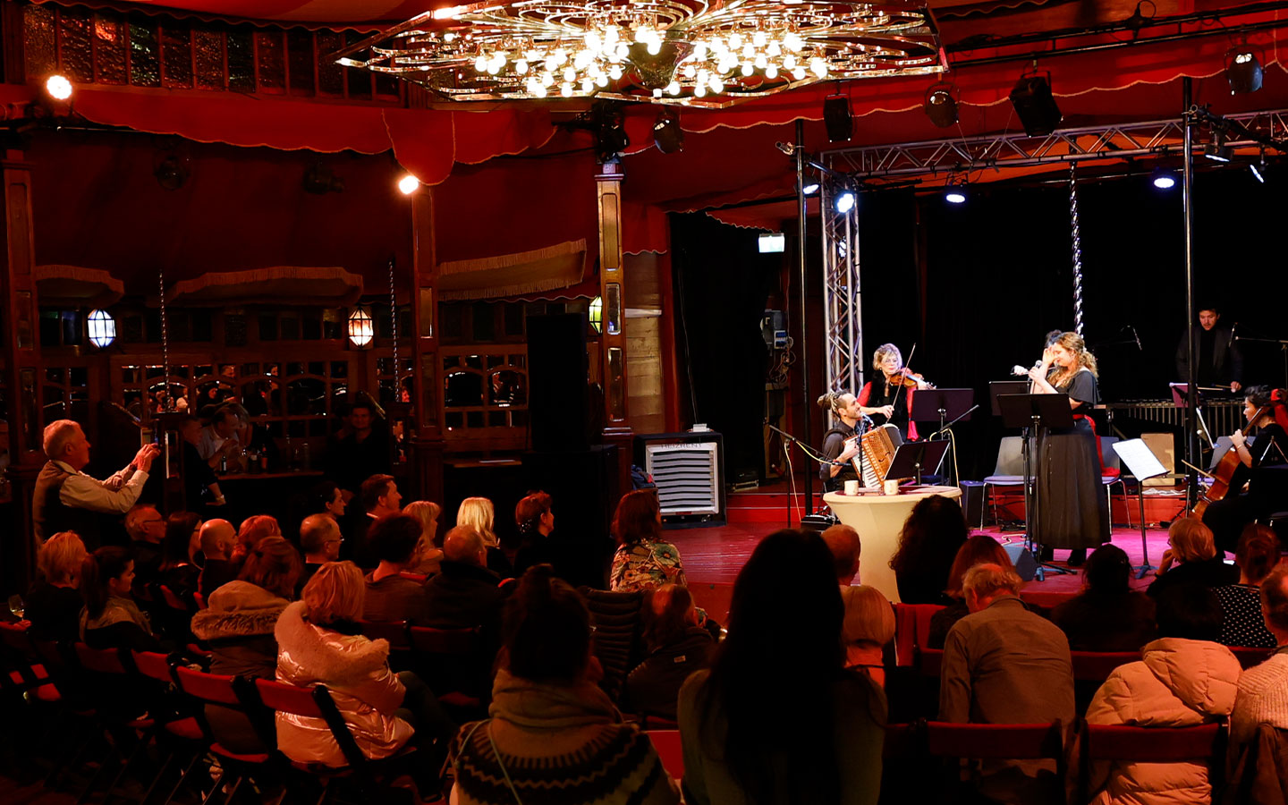 Ein Streichquartett spielt auf einer hell erleuchteten Bühne in einem roten Zelt. Das Publikum sitzt in Stuhlreihen und schaut zu. Ein Kronleuchter hängt darüber, und einige Menschen stehen da und fotografieren oder beobachten die Aufführung.
