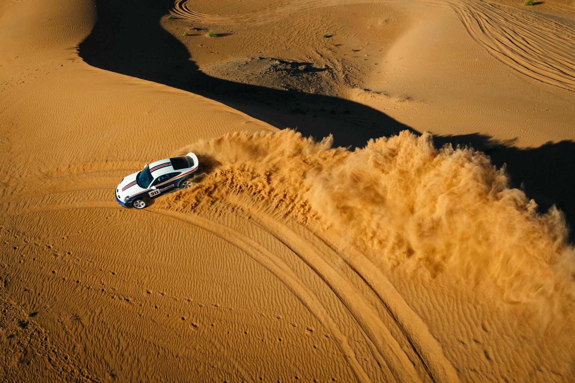 Ein weißes Rallyeauto rast durch die Wüste und wirbelt eine große Sandwolke auf, als es auf den orangefarbenen Dünen unter dem klaren Himmel eine scharfe Kurve macht.