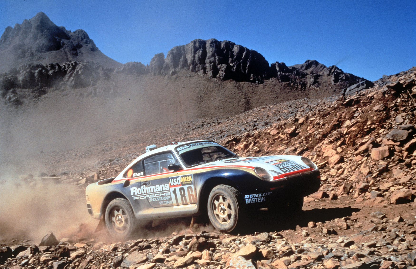 Ein Rallyeauto mit Rothmans-Sponsoring rast über ein felsiges, staubiges Gelände mit Bergen im Hintergrund unter einem klaren blauen Himmel.