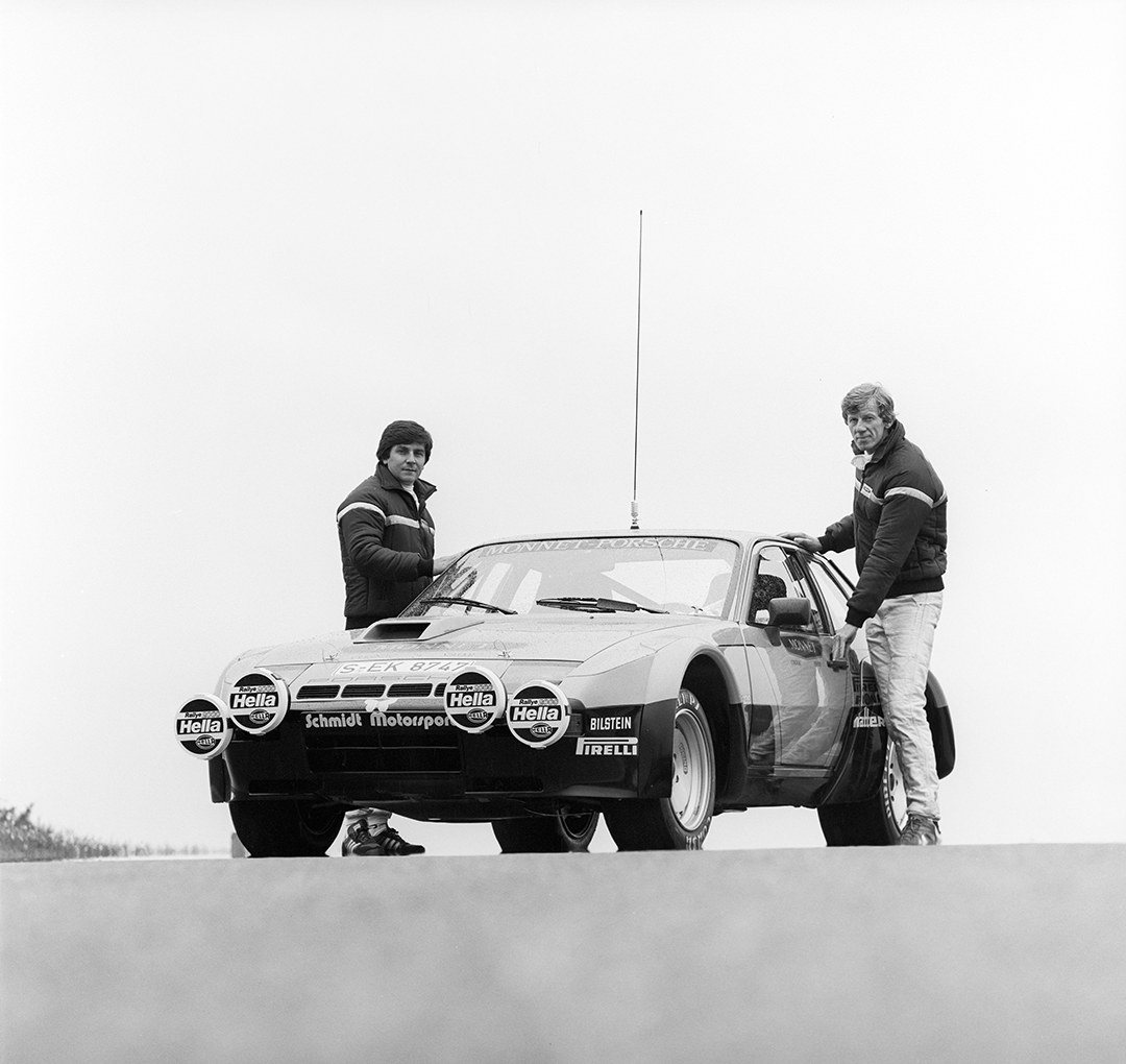 Zwei Männer in Rennanzügen stehen neben einem Oldtimer-Sportwagen mit Rallye-Aufklebern und vier Hella-Scheinwerfern an der Front, fotografiert in Schwarz-Weiß vor einem schlichten Hintergrund.